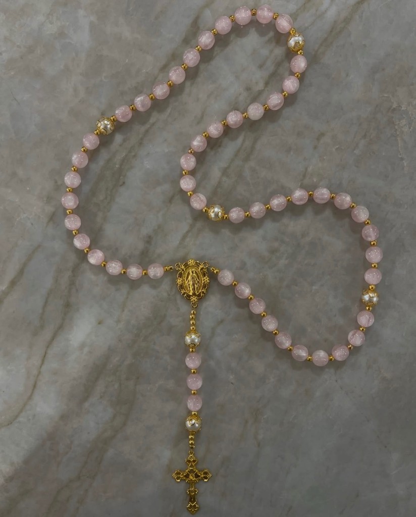 A pale pink Heavenly Rosaries piece with pearl accents and an ornate gold crucifix, photographed on marble.