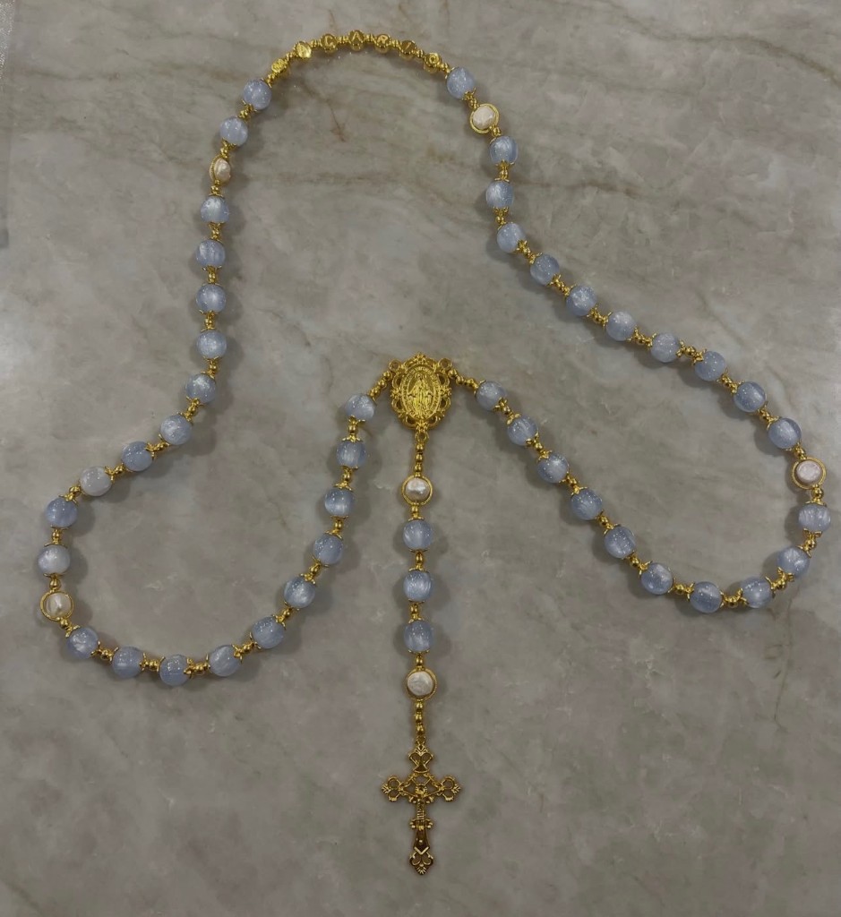 Light blue cat-eye rosary with pearl accents, gold letter beads spelling a name, and an ornate gold crucifix.